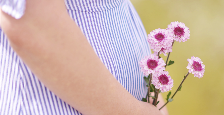 You feel exhausted. And nauseous. But does that mean you're pregnant? Photo of a pregnant belly in a blue dress holding pink flowers, courtesy of Ashton Mullins
